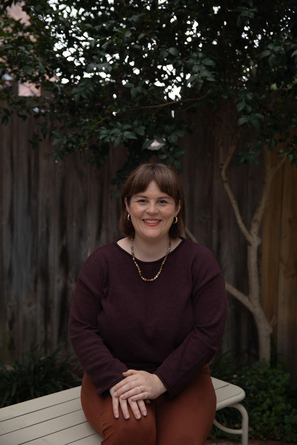 Woman sitting on a bench outdoors with a wooden fence and tree in the background