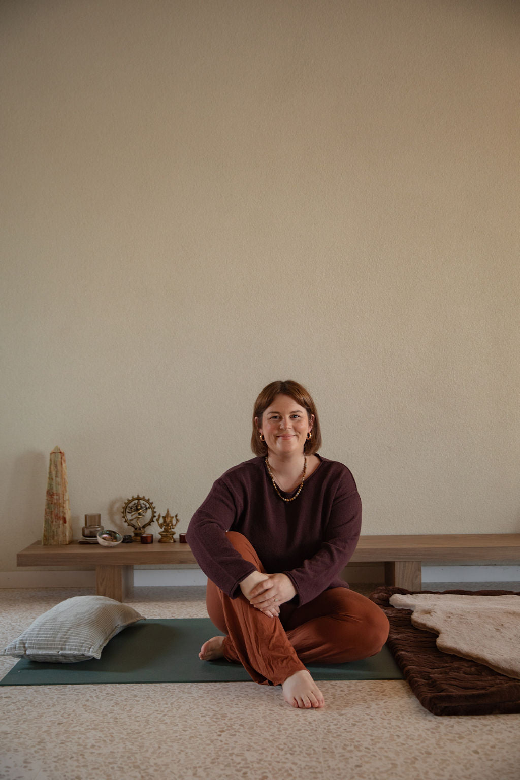Woman sitting on a yoga mat in a minimalistic room with a wooden bench and decorative items.